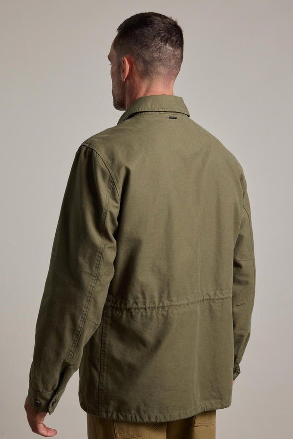 A man with short dark hair, seen from behind, wears the Barkers Nowell Field Jacket in olive green cotton with a collar and buttoned cuffs. The plain, neutral background emphasizes the jacket’s classic utilitarian style.