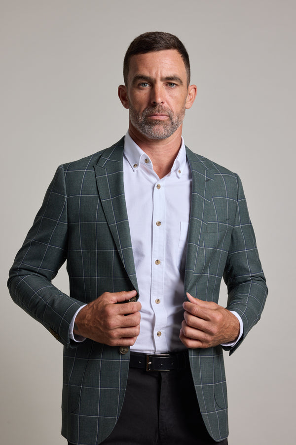 A man with short dark hair and a trimmed beard faces forward against a light gray background, wearing a white button-up shirt and the Barkers Nathaniel Check Blazer, holding the sides of his stylish wool blend blazer.