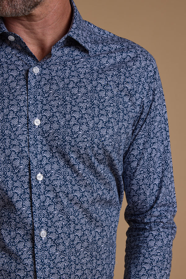 A close-up of a man wearing the Barkers Watkins Print Business Shirt, featuring an intricate white floral pattern on blue Eco Easy Care fabric. Only his lower face, neck, and torso are visible against a solid tan background.
