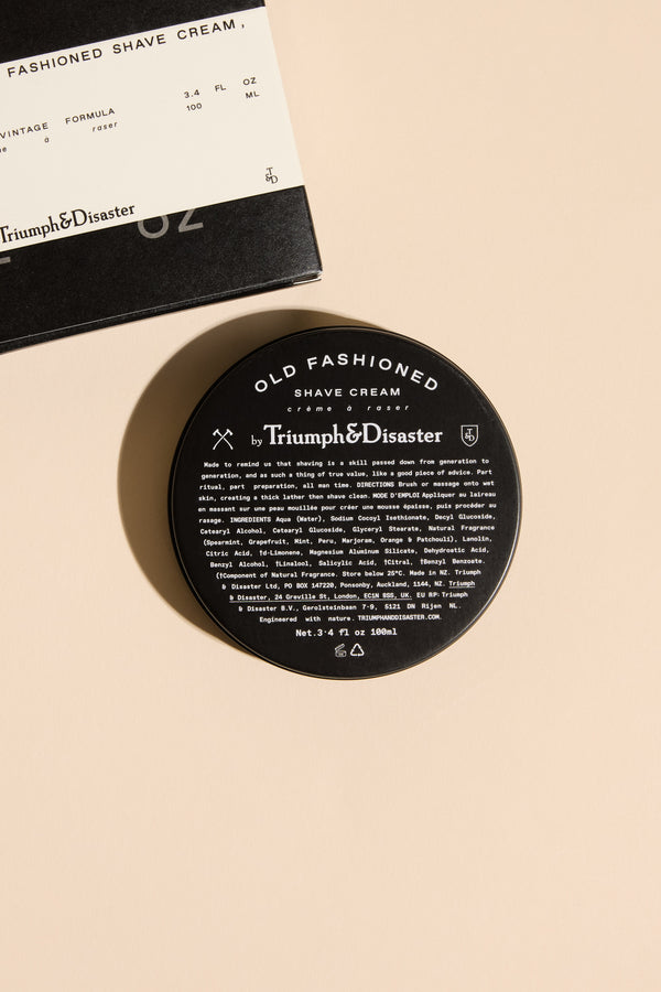 A black jar of Triumph & Disaster Old Fashioned Shave Cream sits on a beige surface beside its matching box, both featuring white text that highlights the traditional shaving style.