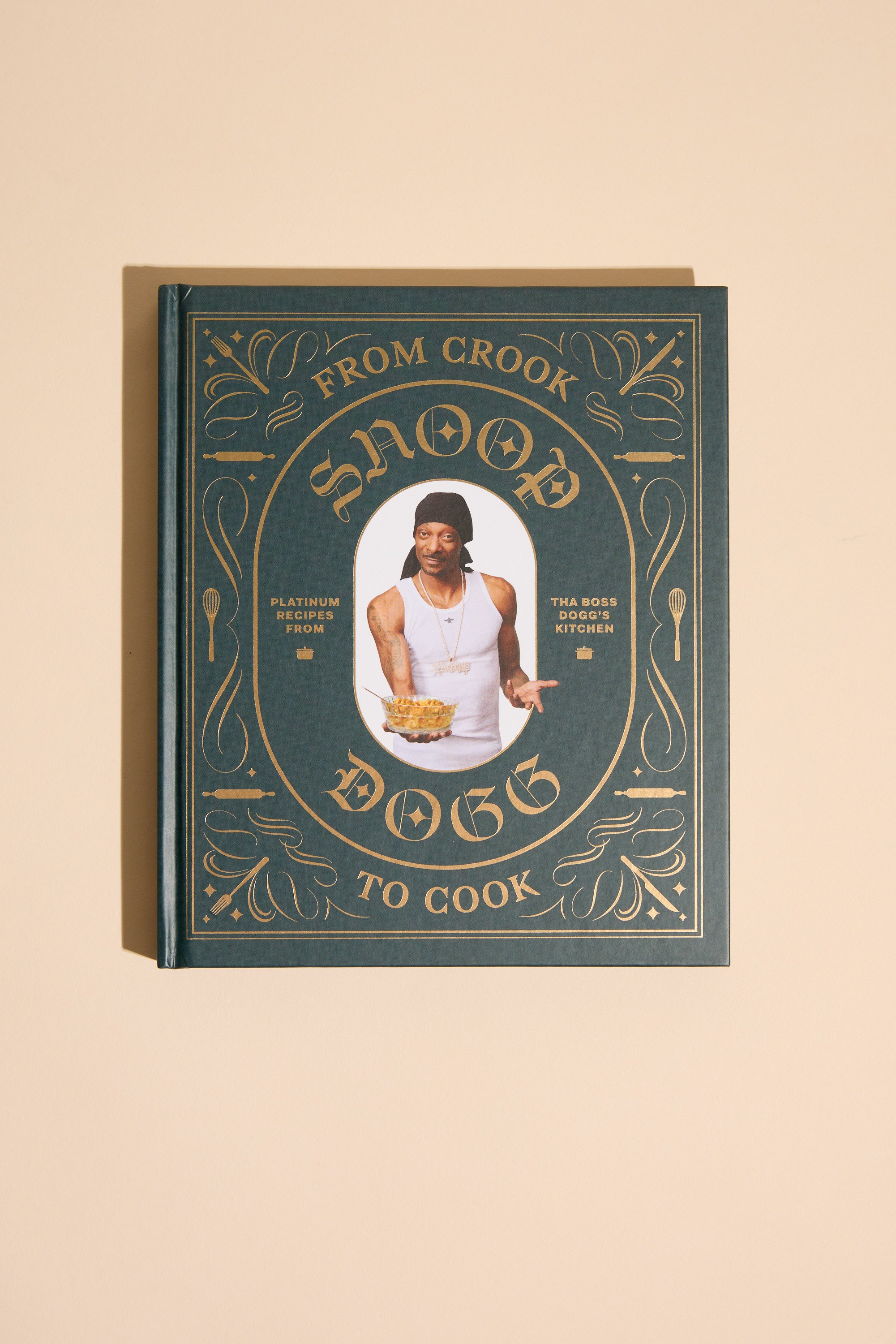 From Crook to Cook by Other is a hardcover cookbook with gold ornate designs on a dark green cover, featuring Snoop Dogg on the front and offering his collection of cannabis-free recipes.