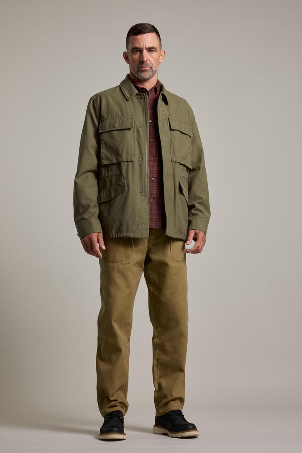 A man stands facing forward against a plain light-gray background, wearing Barkers' Nowell Field Jacket in olive green over a maroon button-up shirt, paired with khaki pants and black shoes.