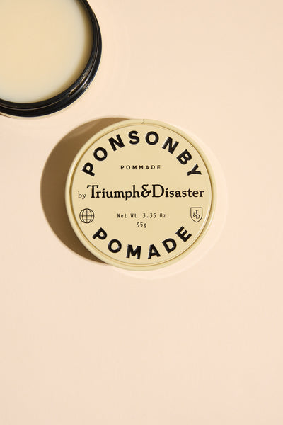 A tin of Triumph & Disaster Ponsonby Pomade, a natural hair pomade by Triumph & Disaster, sits on a light beige surface with its lid off, revealing the product and its bold black label with a minimalist design.