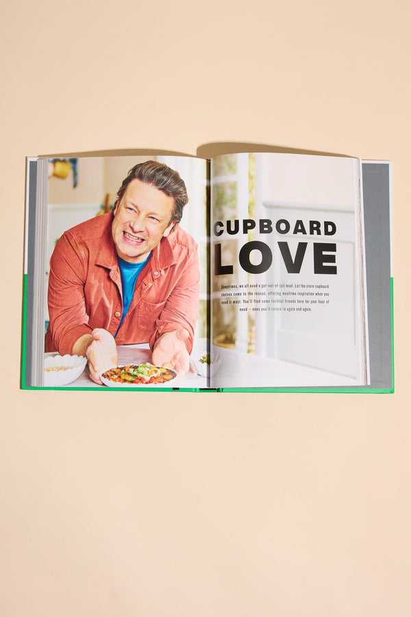 An open Simply Jamie cookbook from Other shows a smiling man in an orange shirt holding a bowl of food, with “CUPBOARD LOVE” as the heading on the right page above a paragraph about easy recipes.