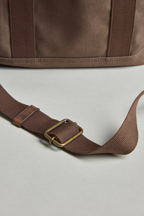 The Barkers Foxton Canvas Convertible Tote is shown in close-up—brown cotton canvas with a matching adjustable strap and brass-tone buckle—laid on a neutral surface. Versatile, it seamlessly transitions from tote to backpack for everyday style.