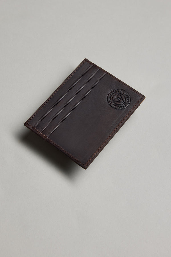 The Barkers Taylor Leather Card Wallet, in dark brown slim leather with three card slots and an embossed circular logo, made from Leather Working Group-certified materials, is shown on a light grey surface.