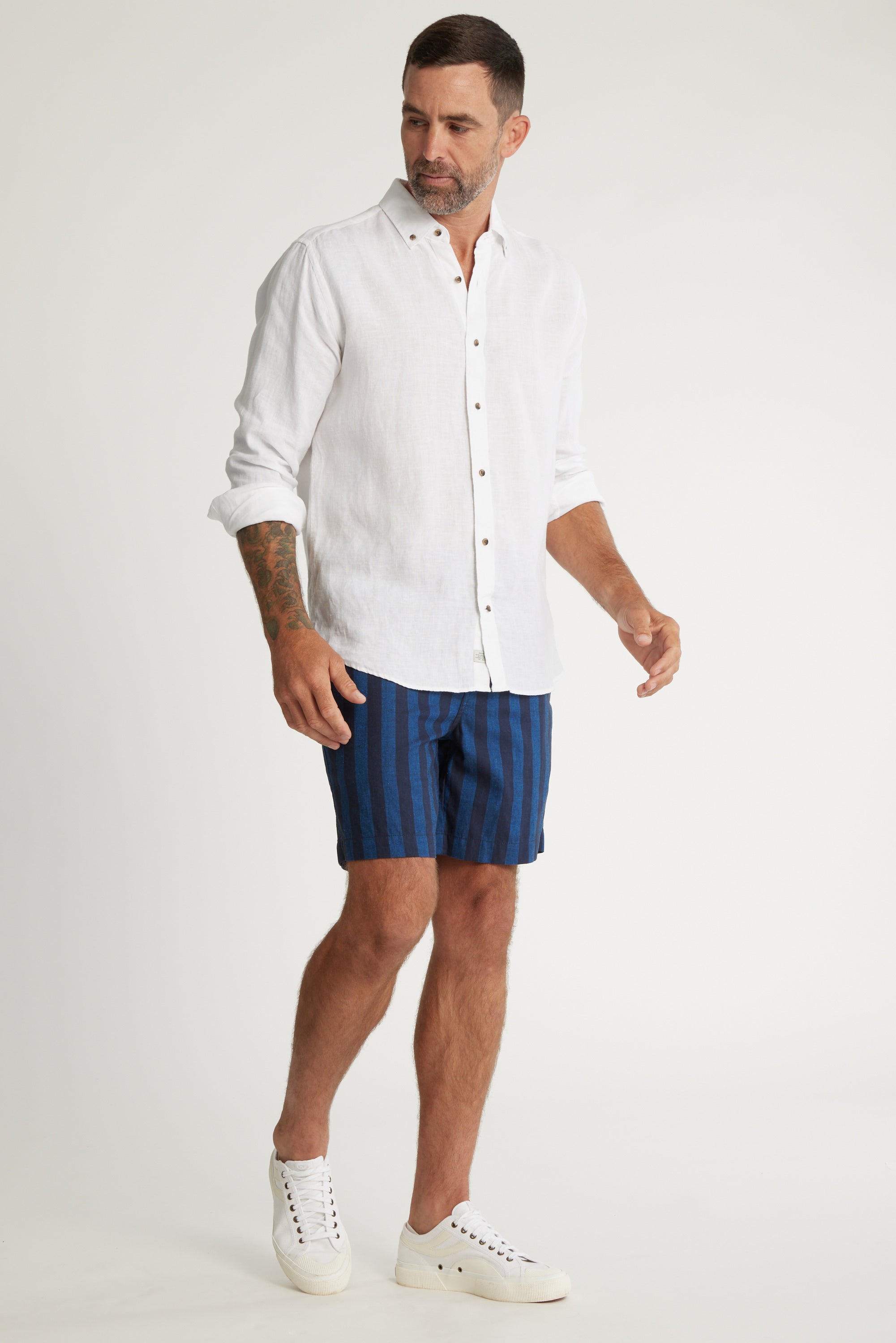 A man with short dark hair and a beard wears a white button-up shirt, Barkers Ashbury Stripe Shorts, and white sneakers. He has tattoos on his left arm and stands against a plain white background.