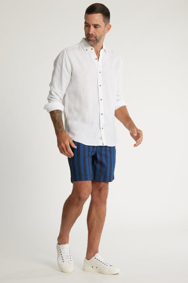 A man with short dark hair and a beard wears a white button-up shirt, Barkers Ashbury Stripe Shorts, and white sneakers. He has tattoos on his left arm and stands against a plain white background.