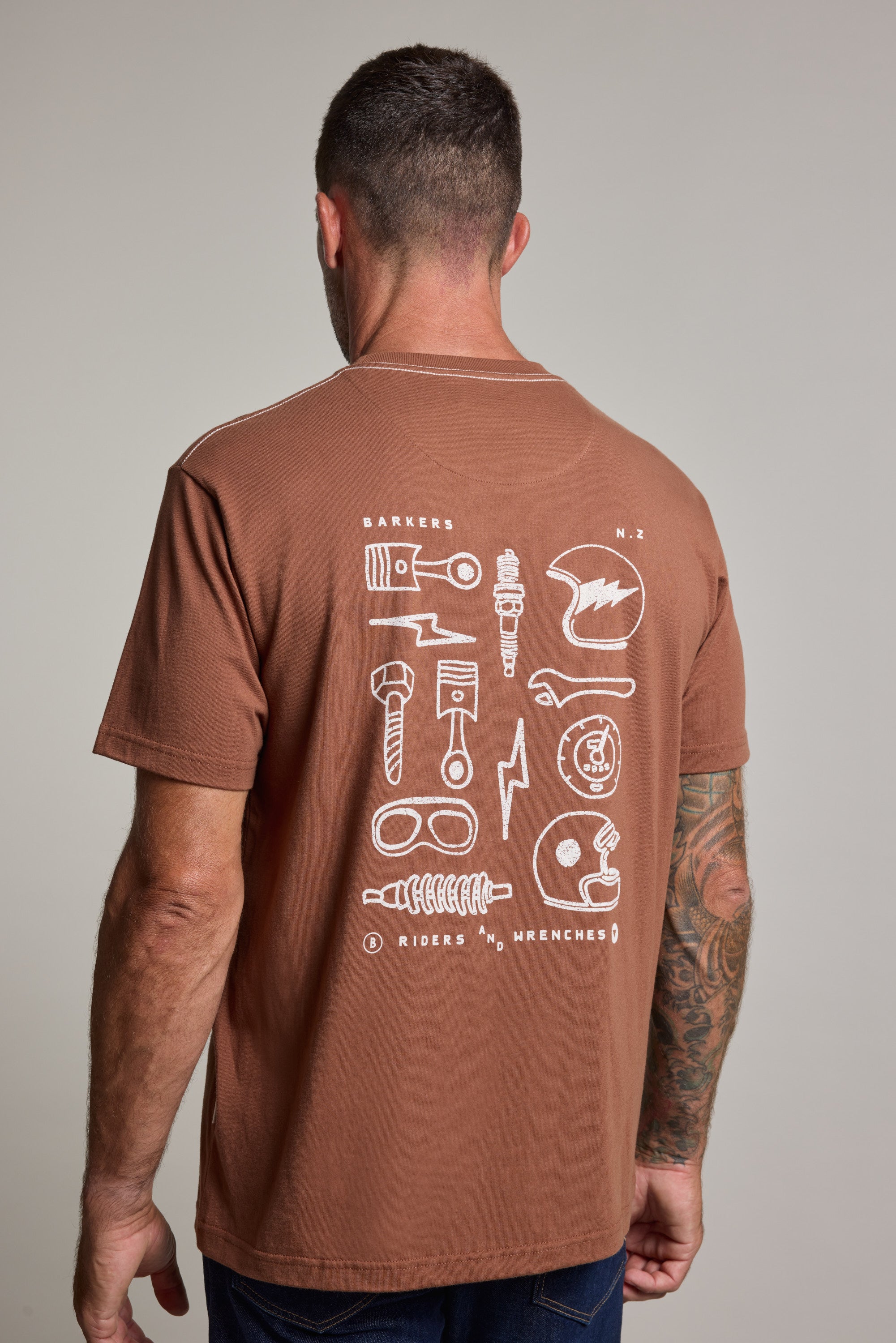 A man with short dark hair and a tattooed right arm wears the Barkers Wrenches Print Tee, a brown organic cotton shirt with white mechanical illustrations and symbols on the back. The photo, taken from behind, highlights the tee’s detailed design.