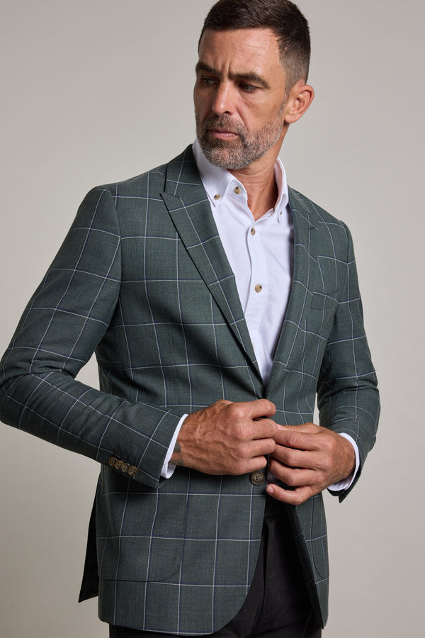 A man with short dark hair and a trimmed beard, wearing the Barkers Nathaniel Check Blazer over a white dress shirt, stands slightly sideways while buttoning his jacket with both hands.