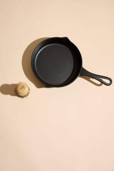 The Ironclad Lil' Legacy Pan by Other, a compact cast iron pan with handle, rests on a beige surface alongside a small round dish scrub brush with natural bristles.