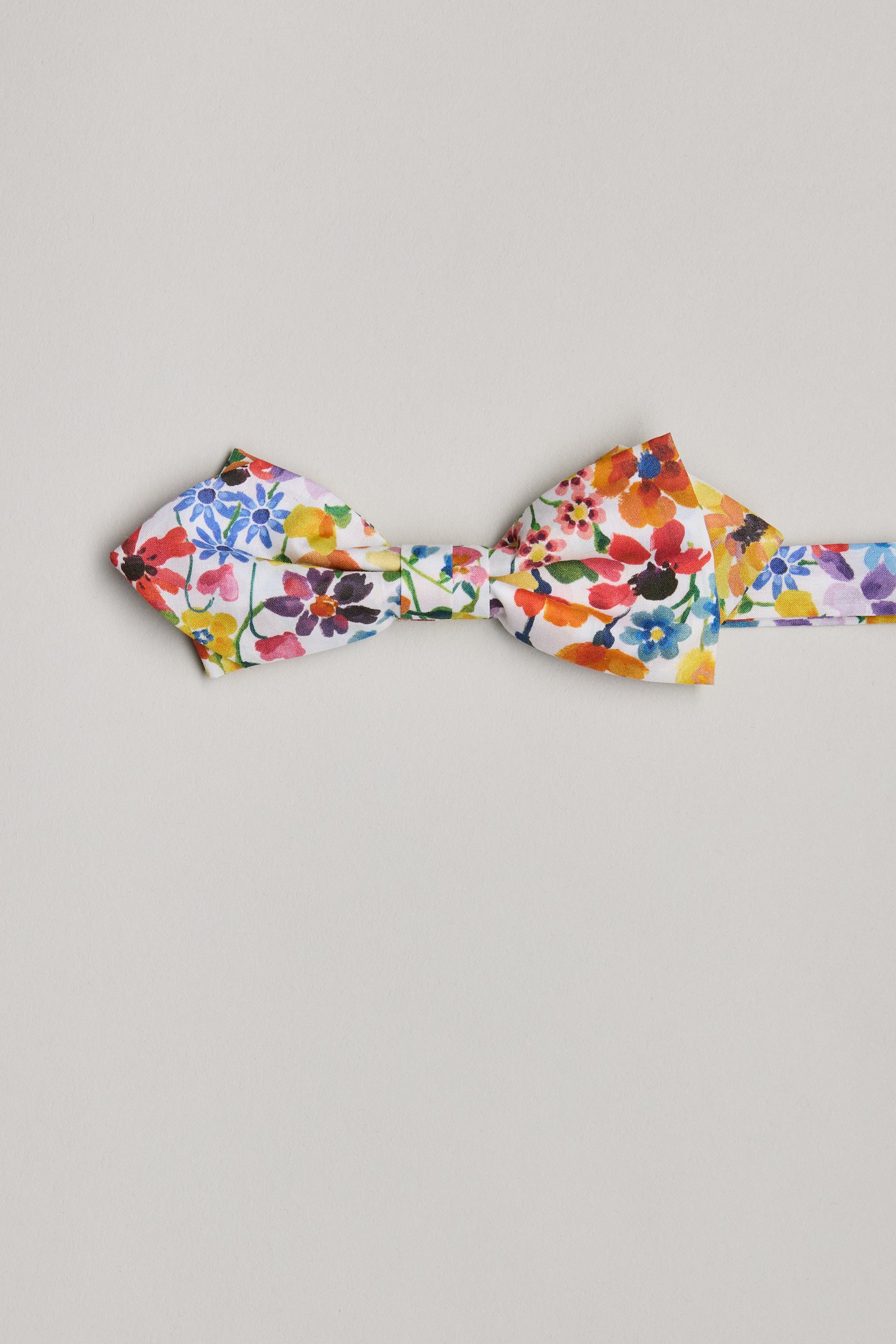 The Dreams of Summer Liberty Bow by Liberty, handmade in New Zealand, showcases a vibrant floral print with small red, orange, blue, and purple flowers on white, beautifully set against a plain light gray background.