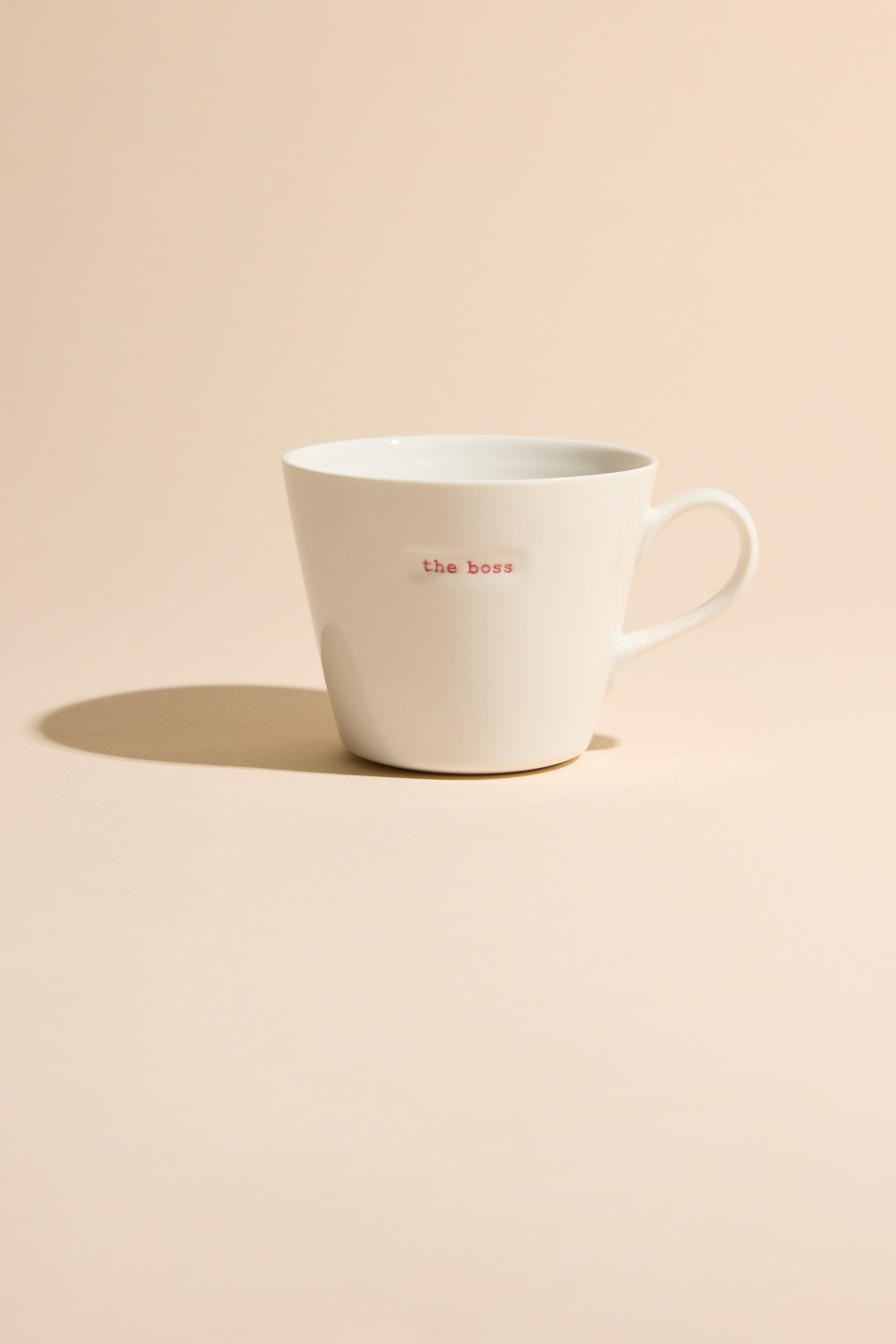 The Bucket Mug - The Boss by Other is a white ceramic mug with "the boss" in small red letters, shown on a light beige surface and matching background, casting a soft shadow to the left.