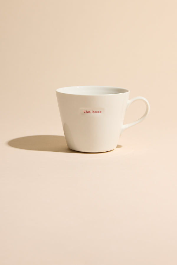 The Bucket Mug - The Boss by Other is a white ceramic mug with "the boss" in small red letters, shown on a light beige surface and matching background, casting a soft shadow to the left.