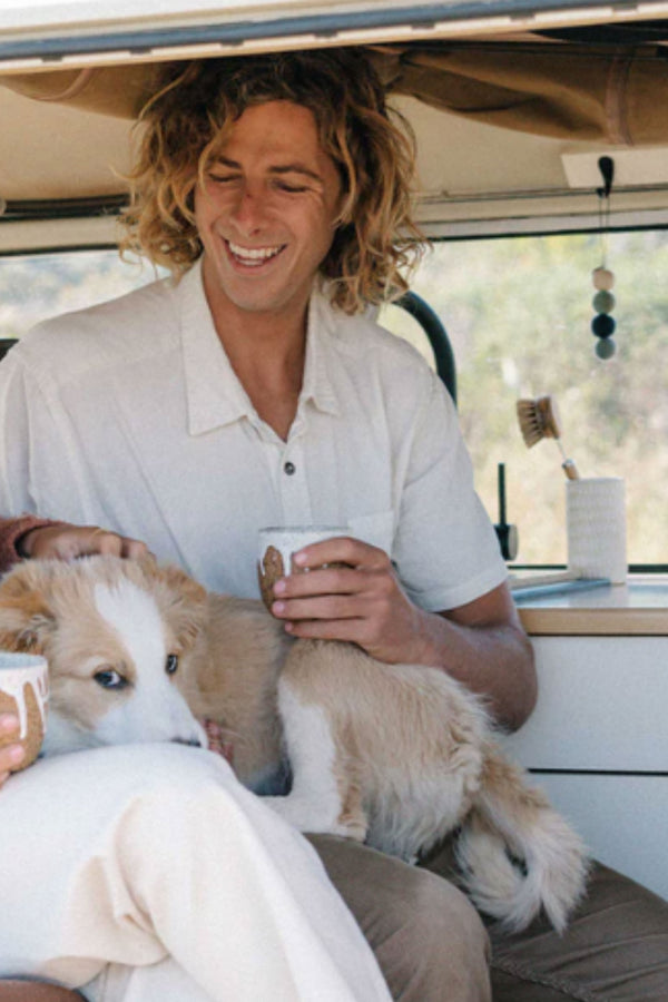 A smiling person with long, wavy hair sits in a vehicle, petting a fluffy brown and white dog on their lap. "Smelly Balls: Cove / Coastal Drift" by Other adds freshness to the cozy scene as cups are held and shared.