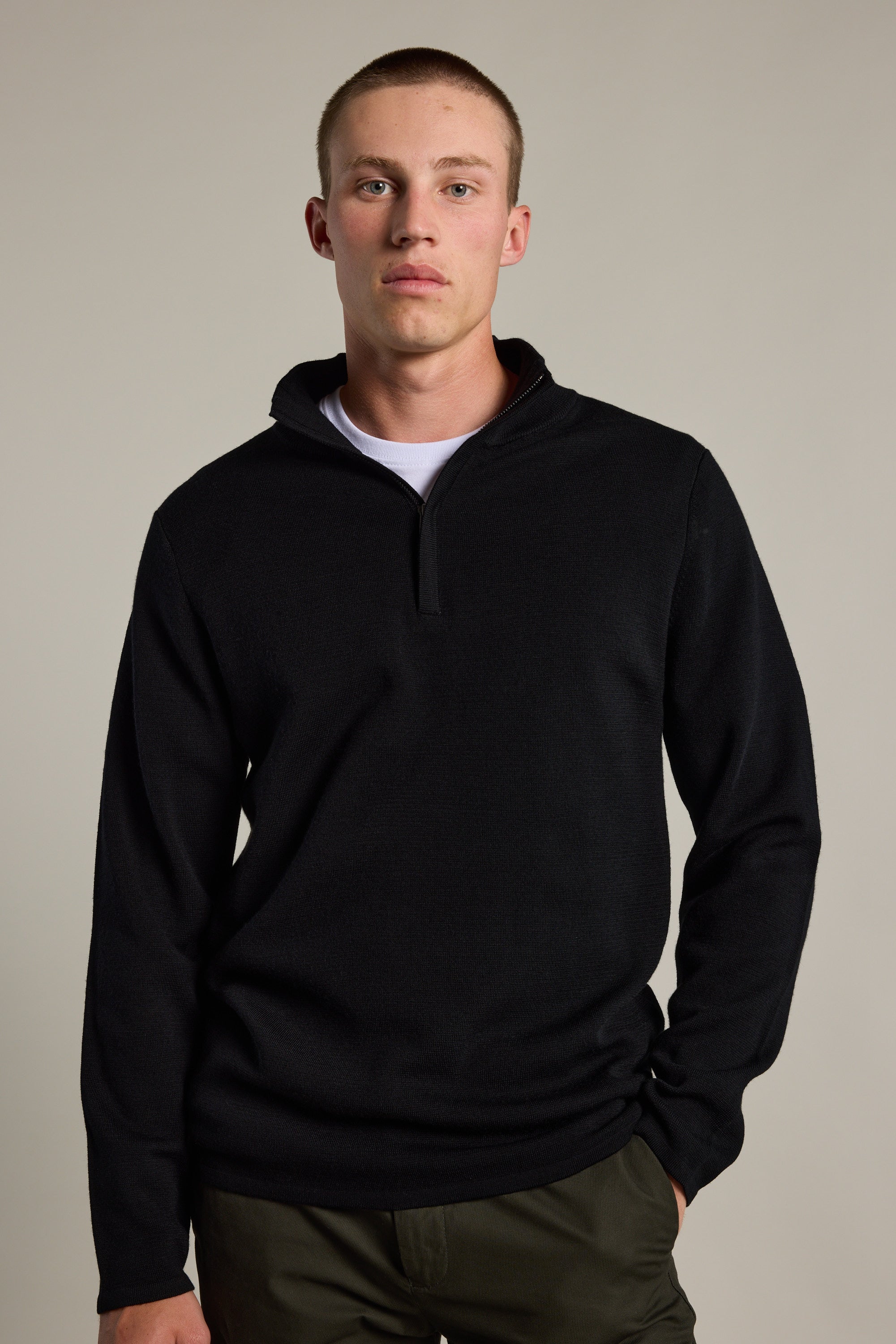 A young man with short hair models the Barkers Brandon ZQRX Merino 1/4 Zip sweater in black over a white tee and olive green pants, standing against a plain light background—ideal for layering in cold weather.
