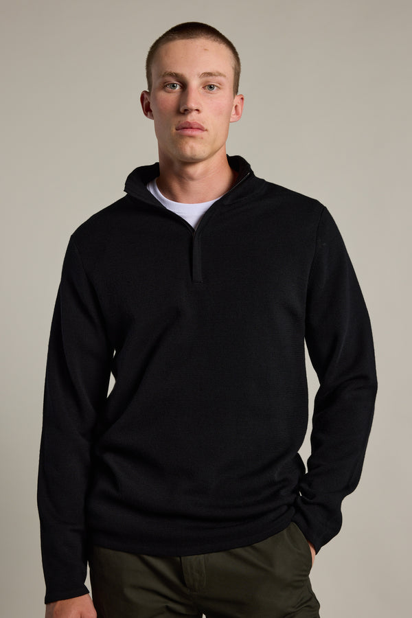 A young man with short hair models the Barkers Brandon ZQRX Merino 1/4 Zip sweater in black over a white tee and olive green pants, standing against a plain light background—ideal for layering in cold weather.