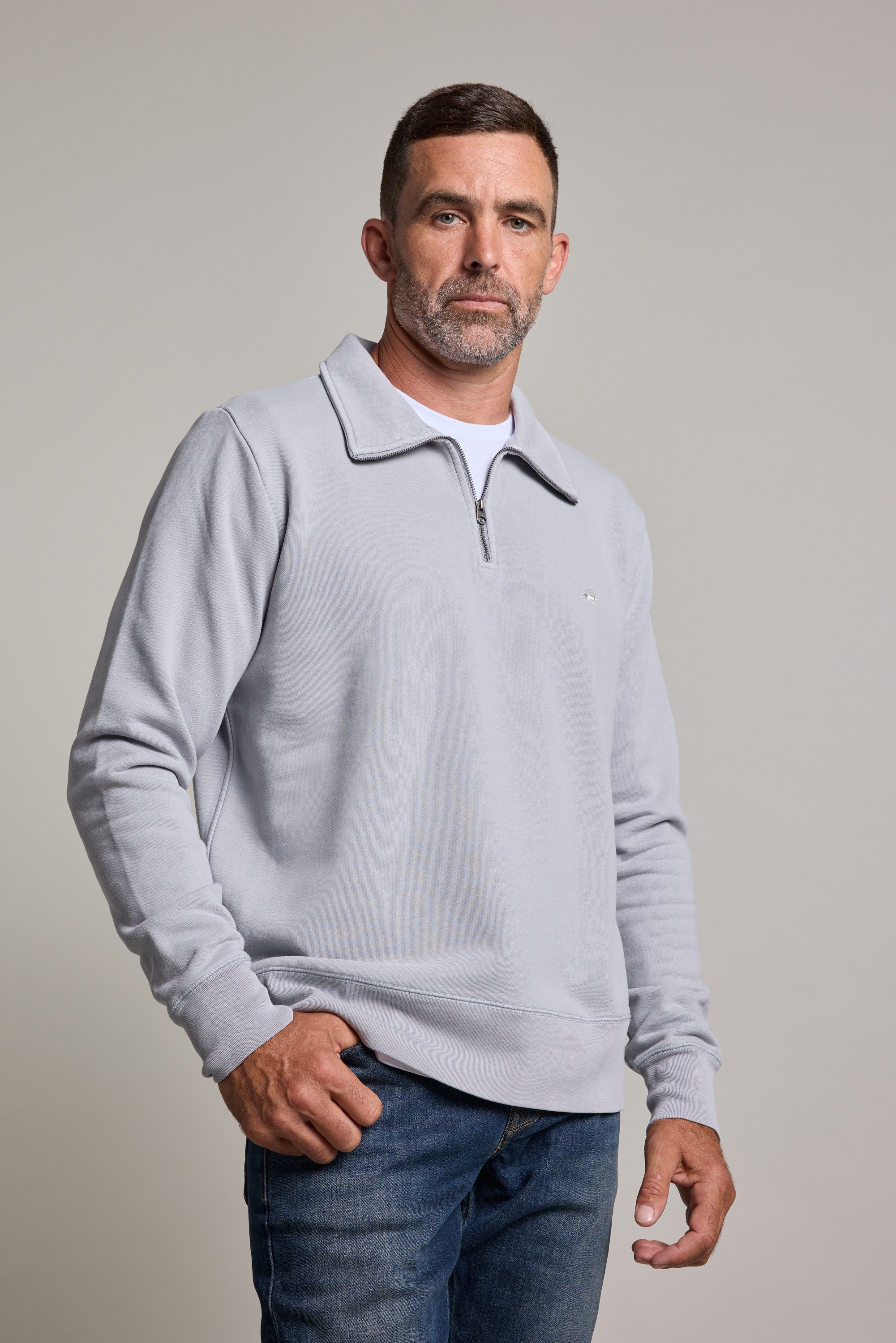 A man with short dark hair and a beard wears the Barkers Galway GD 1/4 Zip in light gray over a white shirt and blue jeans, standing against a plain light background and looking calmly at the camera.
