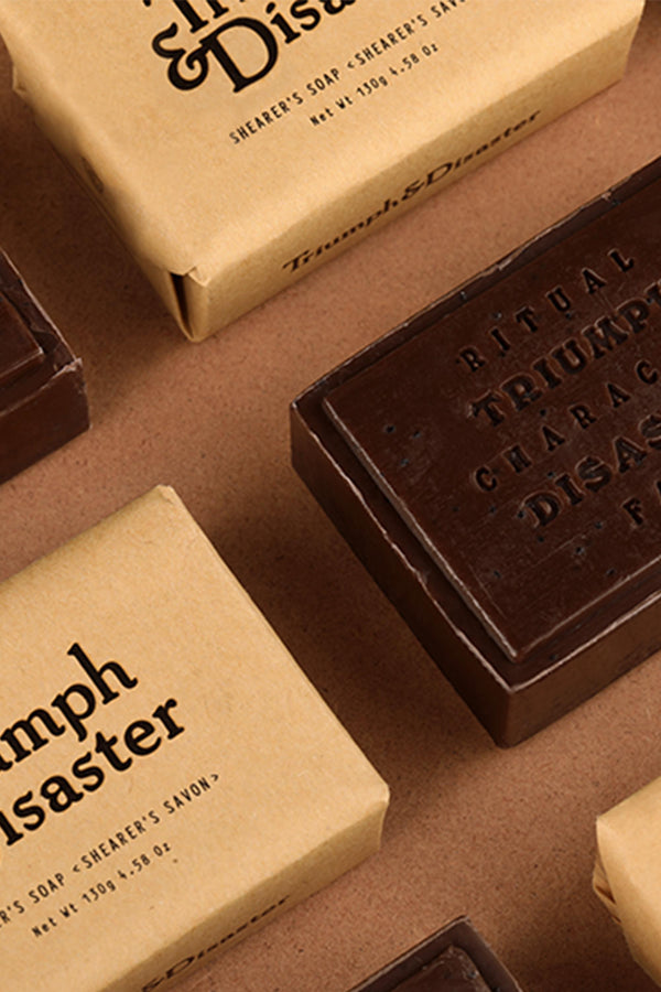 Brown rectangular Triumph & Disaster Shearers Soap bars and tan cardboard boxes are arranged on a brown surface. Each bar is labeled with the brand and product name in bold black letters, highlighting their gentle, exfoliating formula.
