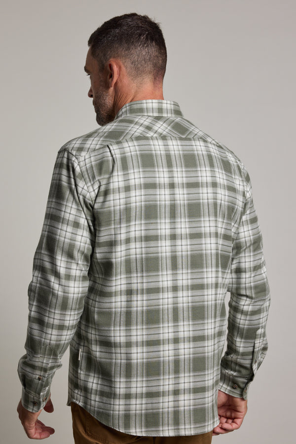 A man stands with his back to the camera, wearing a green check Barkers Paxtonheck Shirt in a classic fit and brown pants, set against a plain light gray background.