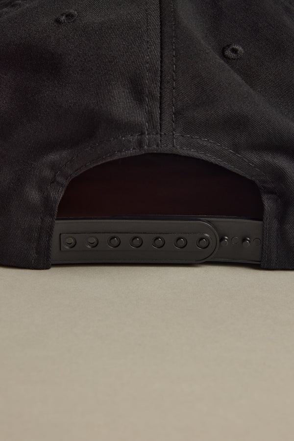 Close-up of the back of the Brixton Payment MP Snapback by Brixton, showing its black unstructured 5-panel design and adjustable plastic snap closure, resting on a light beige surface.