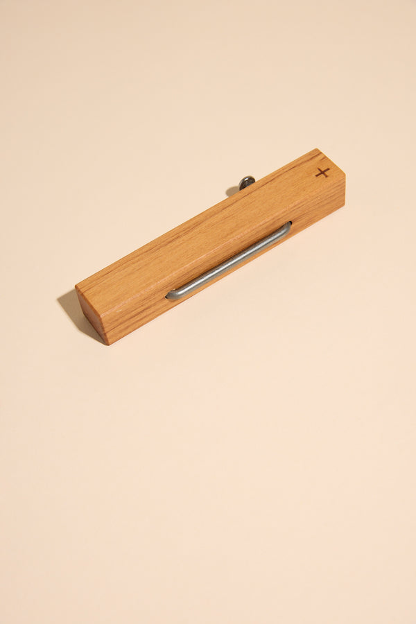 The Other Rimu & Nail Bottle Opener features handcrafted wood, a metal clip, and an engraved plus sign on one end, shown resting on a simple beige surface.