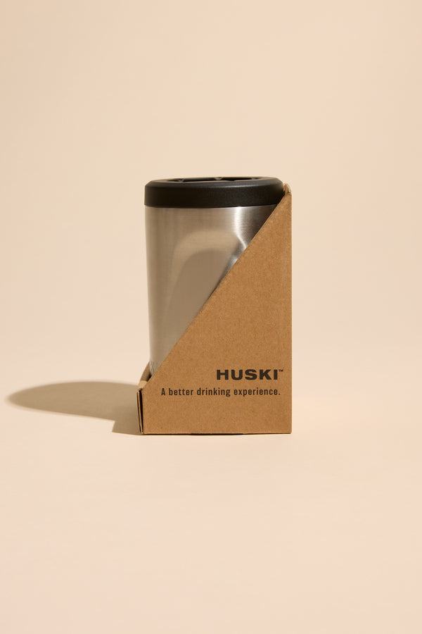 A Huski Beer Cooler by Huski holds a stainless steel tumbler with a black lid, set against a beige background, offering an enhanced drinking experience.