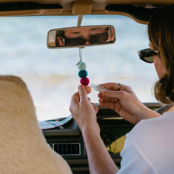 A person wearing sunglasses sits in a car with colorful Smelly Balls: Roadie / Tobacco Vanilla by Other, a reusable car air freshener, hanging from the rearview mirror and holding a small object, with the ocean visible through the windshield.