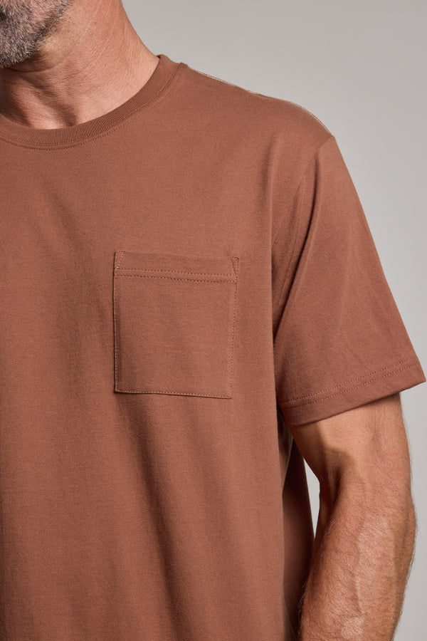 A close-up of a person wearing the Barkers Wrenches Print Tee, a brown heavyweight t-shirt featuring a small chest pocket on the left; only the lower face, neck, and upper body are visible.