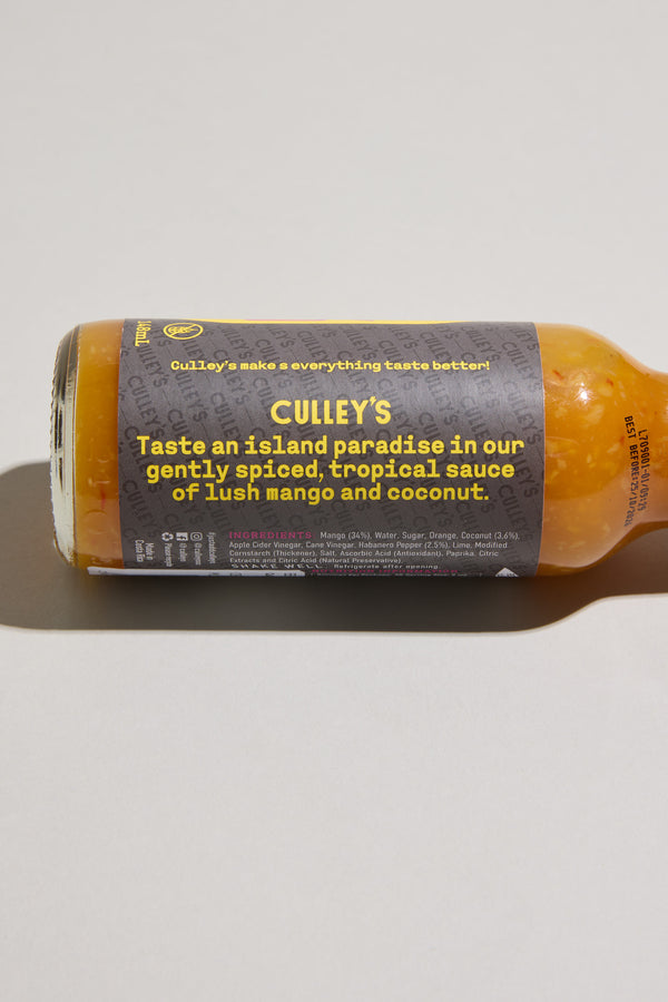 A bottle of Culley's Dreamy Mango-Coco Hot Sauce lies on its side, showing a label that highlights its gently spiced mango coconut flavor with a touch of habanero. The sauce inside is bright orange.