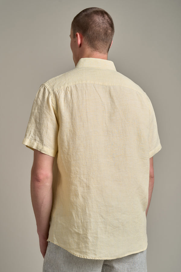 A man faces away from the camera in a light yellow Gentry Linen Shirt by Barkers and light, vertically striped shorts, set against a beige background—ideal for any summer wardrobe.