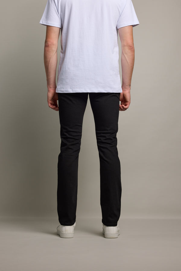 A person stands facing away from the camera, wearing a white T-shirt, Barkers Rambler Denim Slim Jean, and white shoes against a plain light-colored background.