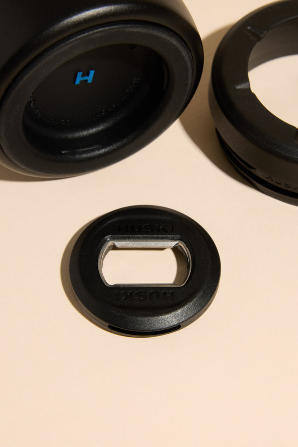 A close-up of black circular objects on a beige surface, including stainless steel components and a Huski-branded bottle opener, designed for use with the Huski Beer Cooler by Huski.