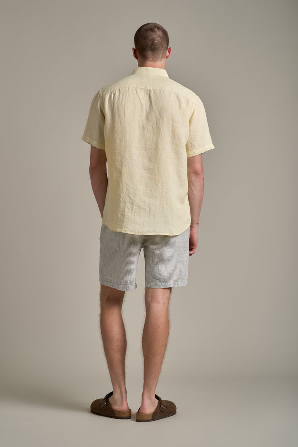 A man faces away from the camera wearing the Barkers Gentry Linen Shirt in pale yellow, paired with light gray striped shorts and brown shoes—an effortlessly cool addition to any summer wardrobe, set against a plain light background.