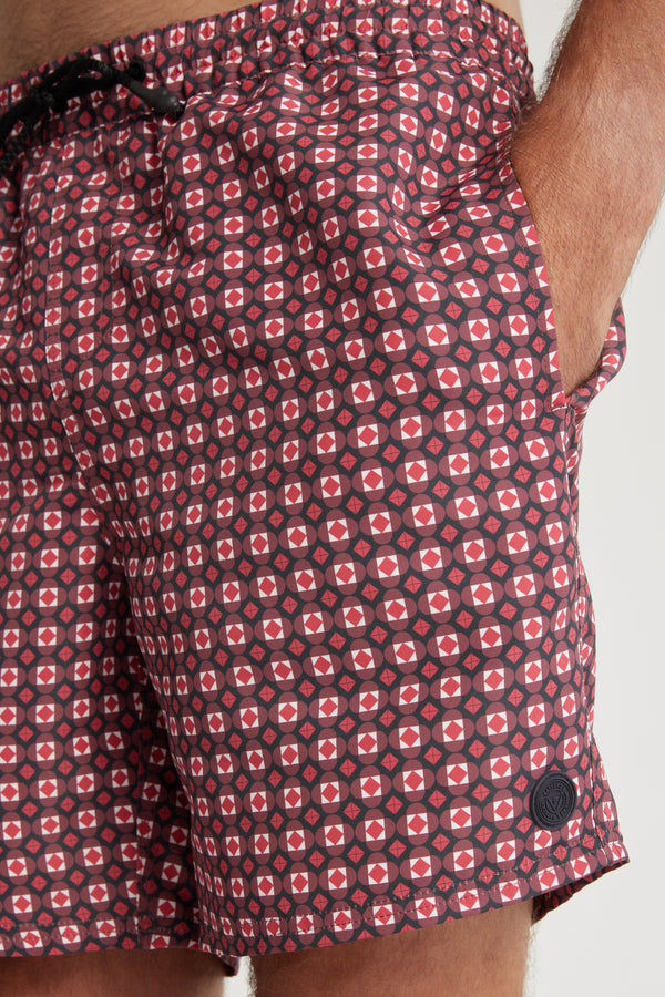 Close-up of a person wearing Barkers Laramie Print Volley shorts, red and black with a geometric pattern, made from recycled polyester. Features include a black drawstring, round logo patch near the hem, and the person’s hand in the pocket.
