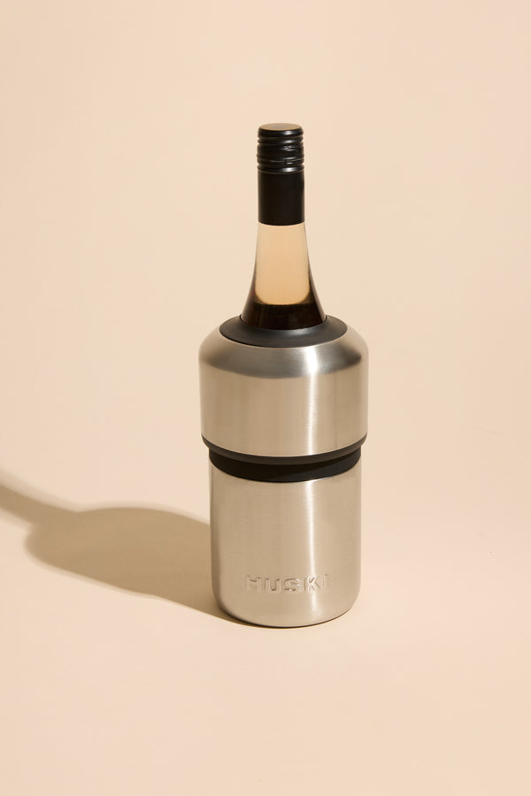 A Huski Wine Cooler by Huski holds a wine bottle on a beige background, casting a soft shadow. This sleek stainless steel cooler keeps your wine cold for hours.