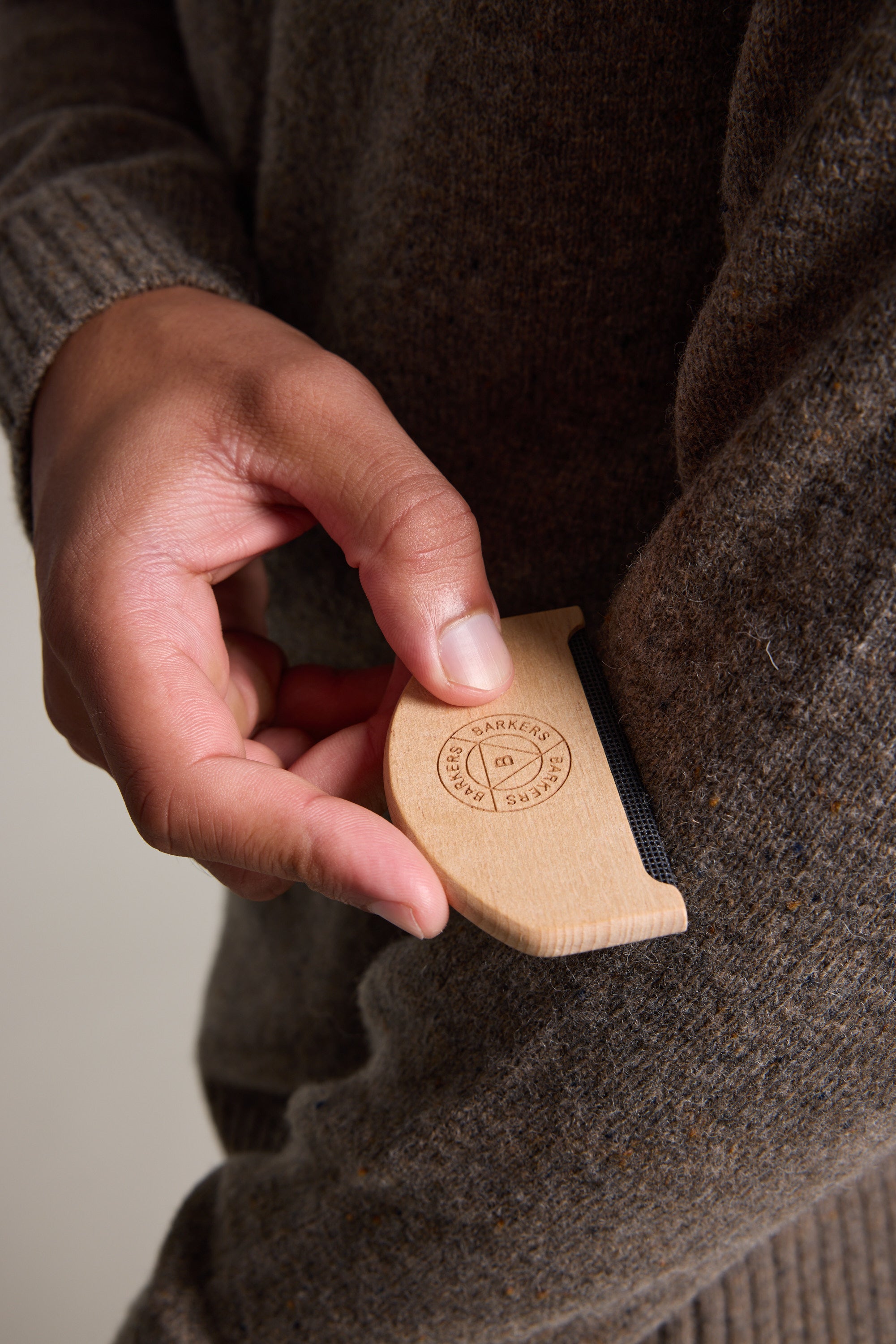A hand uses the Barkers Sweater Comb on a brown wool sweater sleeve, gently removing pills and lint from the knitwear. The comb features an engraved Barkers logo and text.
