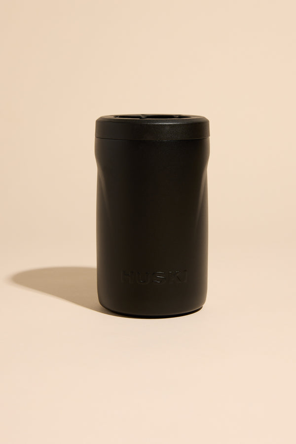 A matte black Huski Beer Cooler by Huski, made of stainless steel with insulation, stands upright on a beige surface. The brand name "HUSKI" is embossed on the lower front, and a soft shadow falls beneath it.