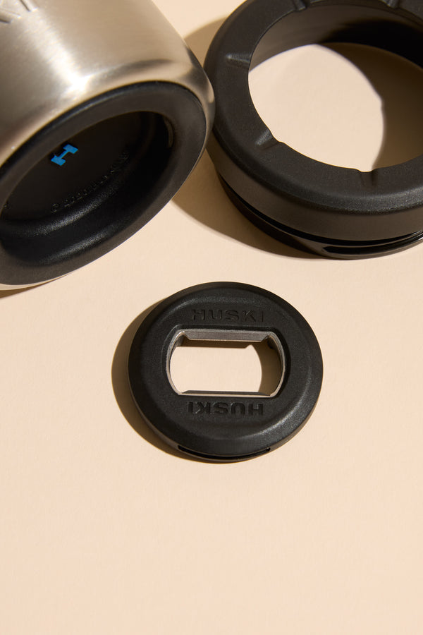 Close-up of a Huski Beer Cooler stainless steel tumbler, part of Huski’s high-performance set, with two black lids—one featuring the Huski-branded bottle opener—displayed on a neutral beige surface.
