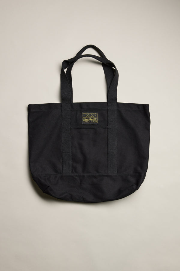 The Barkers Supply Co. Canvas Tote Bag by Barkers features two handles and a small rectangular patch label at the top center. Made from durable black canvas, it's displayed against a plain light background.