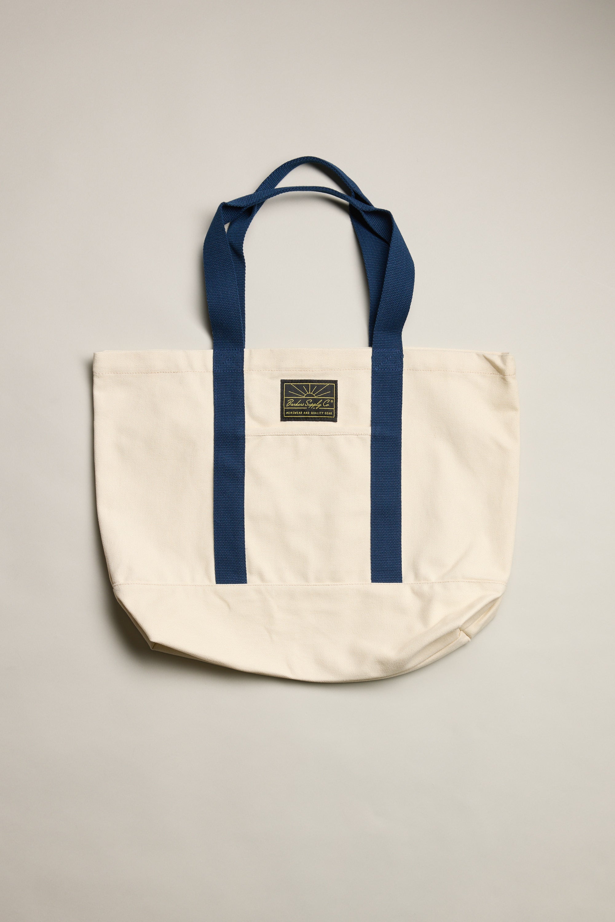 The Barkers Supply Co. Canvas Tote Bag by Barkers is a durable cream canvas tote with two blue handles, matching vertical stripes, and a small rectangular logo patch—perfect for everyday travel use.