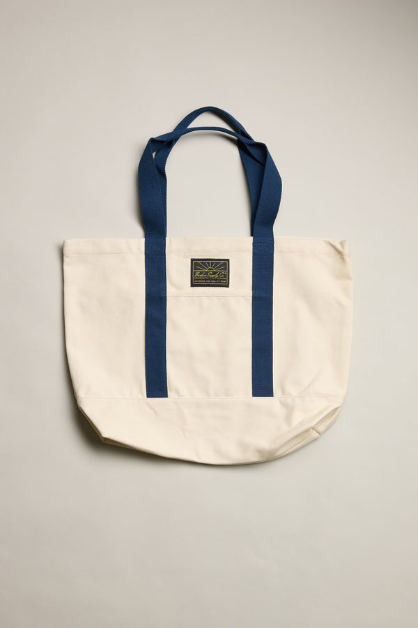 The Barkers Supply Co. Canvas Tote Bag by Barkers is a durable cream canvas tote with two blue handles, matching vertical stripes, and a small rectangular logo patch—perfect for everyday travel use.