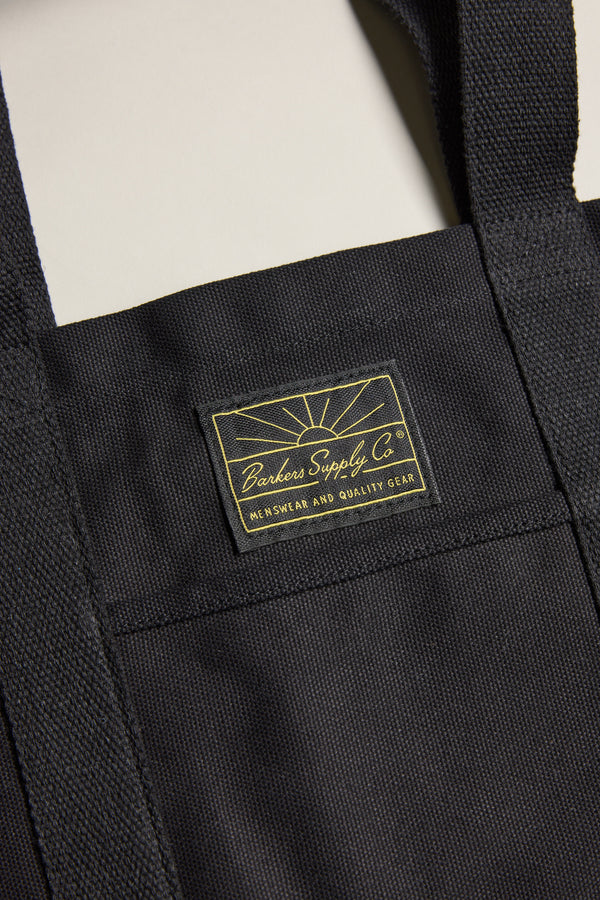 Close-up of the Barkers Supply Co. Canvas Tote Bag by Barkers, made from durable black canvas with sturdy handles, visible stitching, and a sewn label featuring yellow text and a sunrise design.