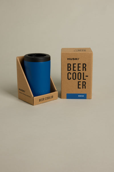 A blue Huski Beer Cooler by Huski in a cardboard holder is next to its brown box packaging labeled "Huski Beer Cooler Ocean Blue," all on a plain, light background.