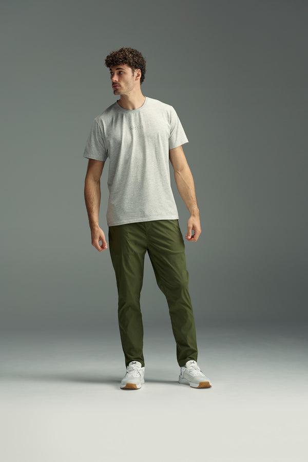 A man with curly hair stands against a plain grey background, wearing a light grey t-shirt, white sneakers, and WearEver’s tapered fit olive green Transit Pant, looking off to the side with a neutral expression.