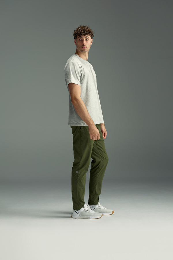 A man with curly hair stands sideways, looking over his shoulder. He wears a light gray T-shirt, white sneakers, and olive green WearEver Transit Pants by WearEver, posed against a plain gray background.