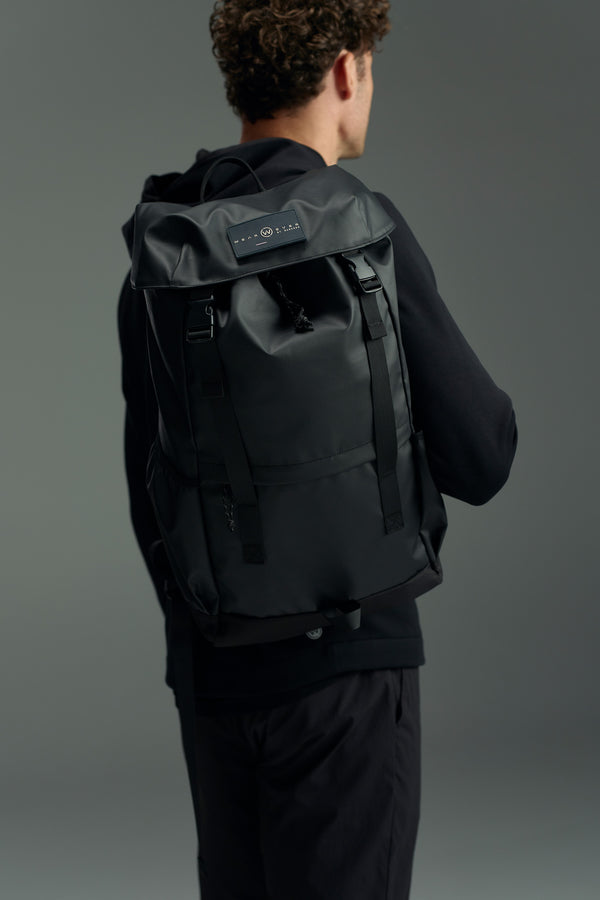 A person with curly brown hair in black attire is seen from behind wearing the WearEver Transit Backpack, which features a rectangular label and water-resistant zips, set against a neutral gray background.
