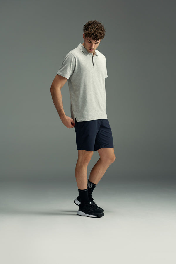 A man stands in a neutral pose wearing the WearEver Outbound Polo by WearEver in light gray, paired with dark navy shorts, black socks, and black athletic shoes against a plain gray background.