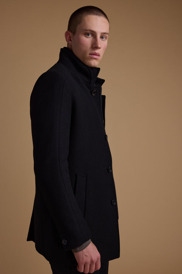 A person with short hair wears the Barkers Raymond Coat, a dark wool blend, standing sideways against a plain beige background and looking at the camera with a neutral expression.