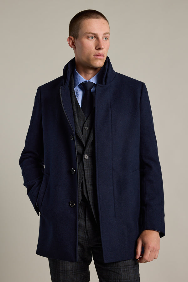 A man wears the Barkers Raymond Coat, a dark blue overcoat, over a gray checkered suit with a blue dress shirt and dark tie. He stands with one hand in his pocket, looking slightly to the side against a plain light background.
